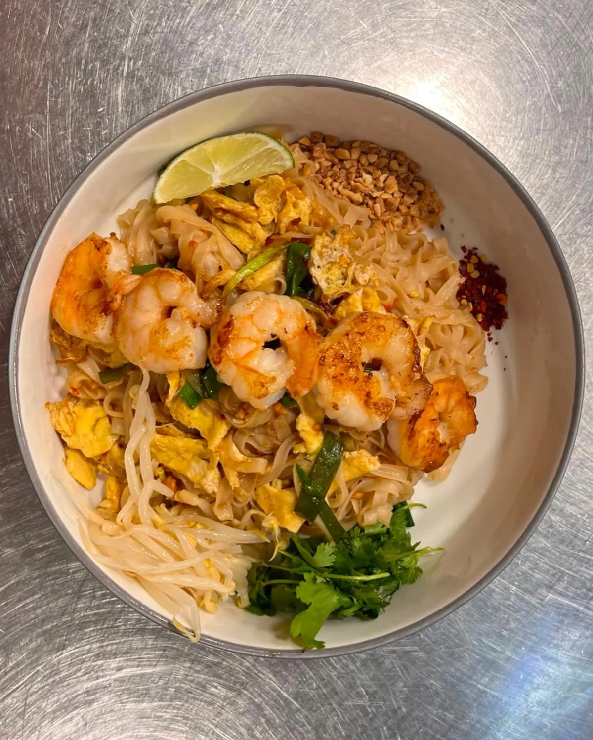 Pad thai: nūdeles ar garnelēm, pupiņu asniem, zaļumiem un laima daiviņu bļodā